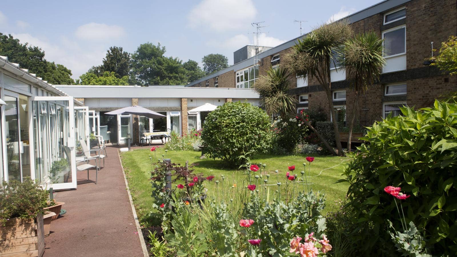 Gardens and flowers at the rear of Kenbrook care home on a sunny day with trees and the doors open to a patio area