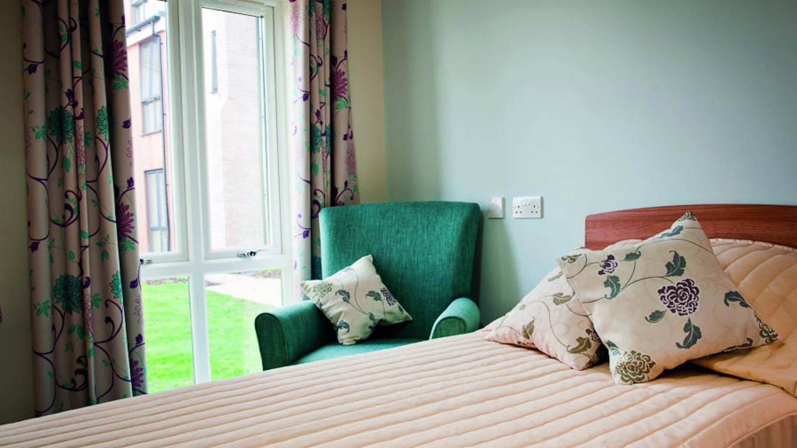 Care home bedroom with a single bed and a comfy chair next to a window and a view outside to communal shared green space