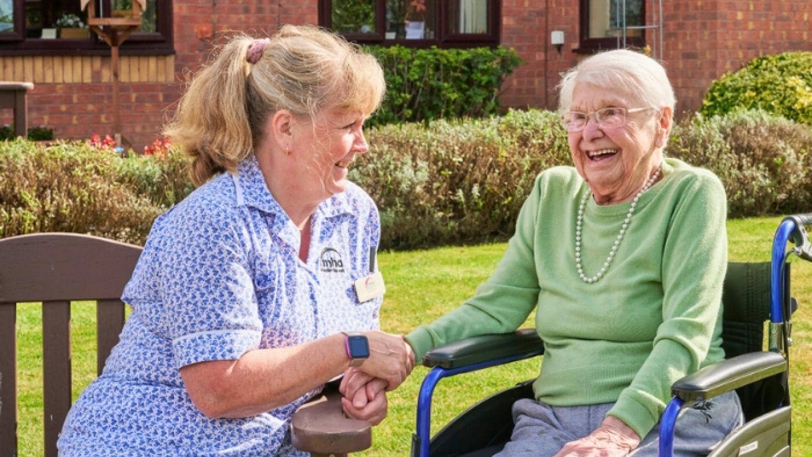 A carer holds the hand of a female resident sitting in a wheelchair in the garden