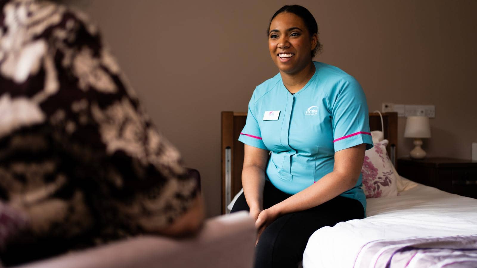 Female care assistant speaking to resident who is out of focus whilst sat on a bed