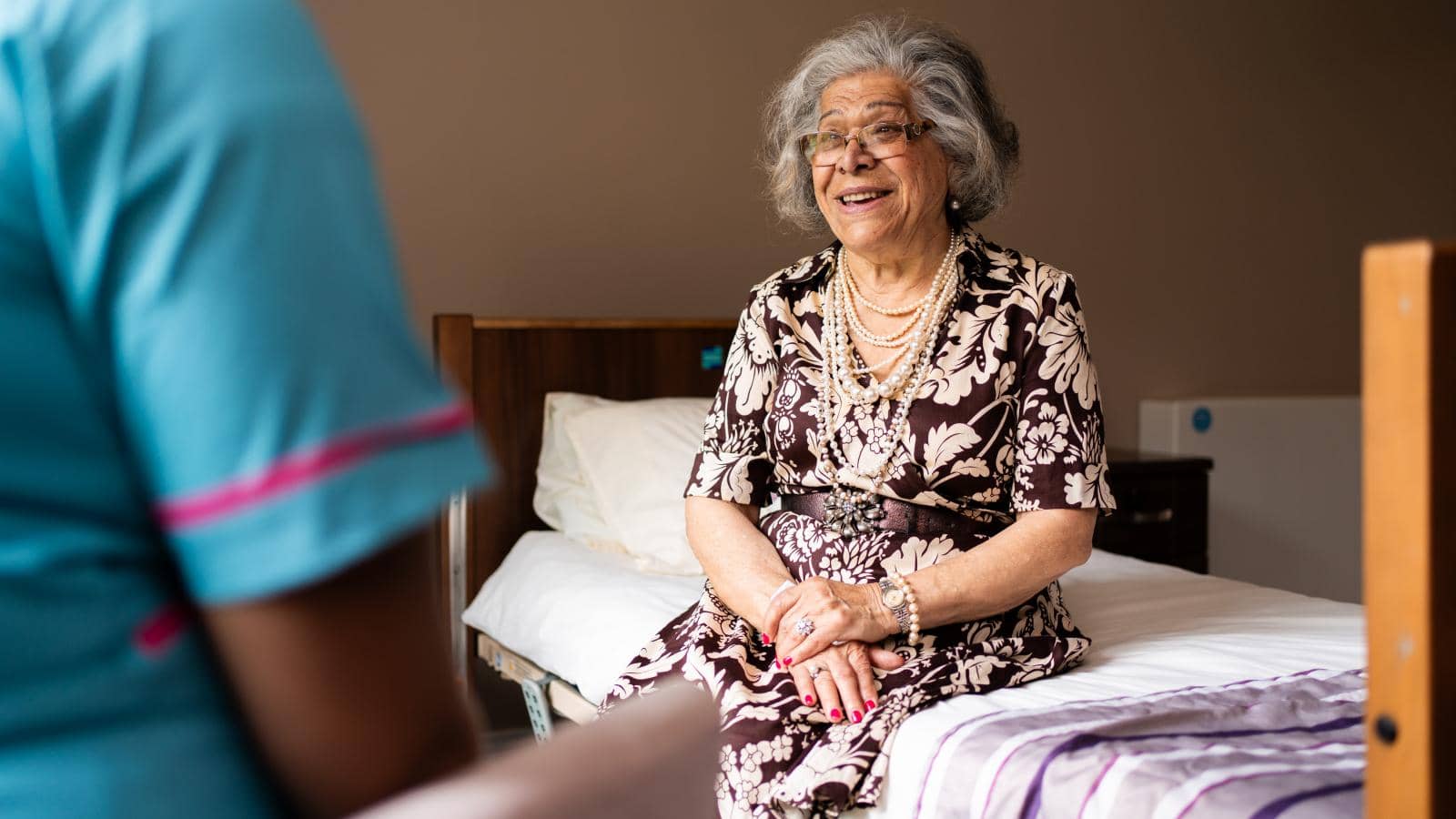 Older female care home resident sat on bed speaking to a care assistant