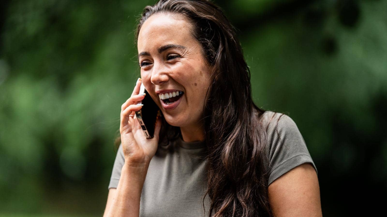 a woman with long, dark hair, smiling and laughing while talking on her phone