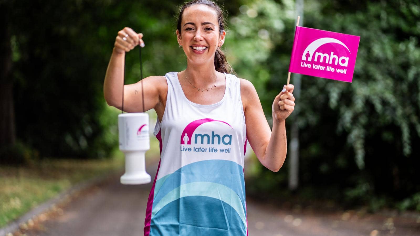 a smiling woman standing on a path surrounded by greenery. She is wearing a sleeveless athletic top with the logo "mha" prominently displayed. In one hand, she holds a collection tin, and in the other, she waves a flag with the same logo