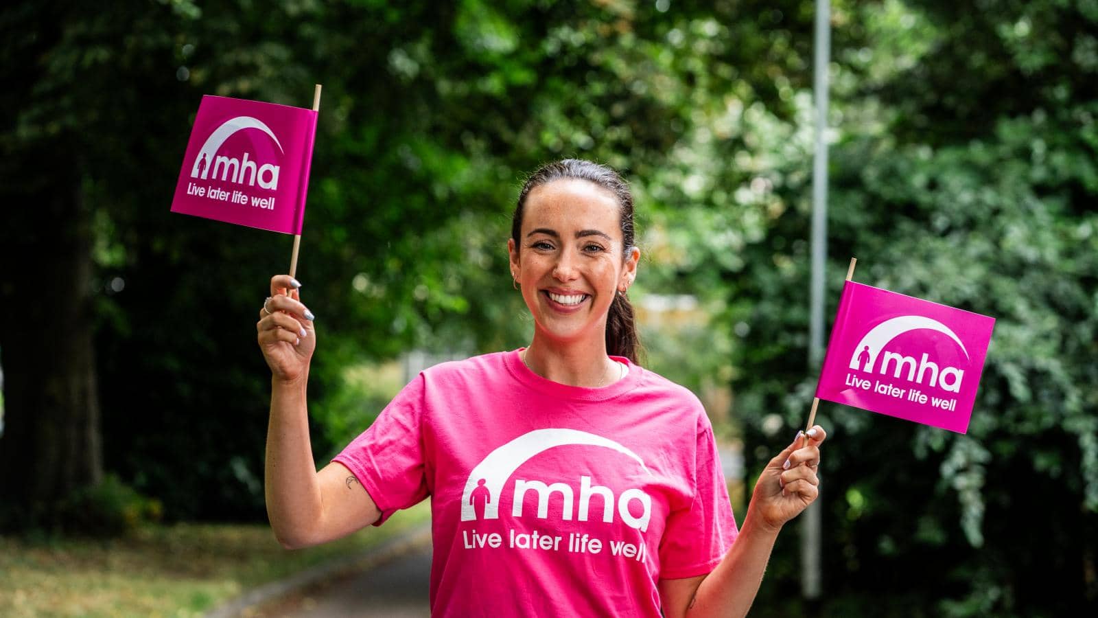 smiling woman holding two small flags, each displaying the logo of "mha" along with the slogan "Live later life well"