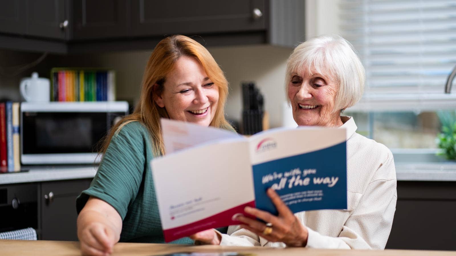 two women sitting at a kitchen table, engaged in a joyful conversation while looking at a brochure or magazine