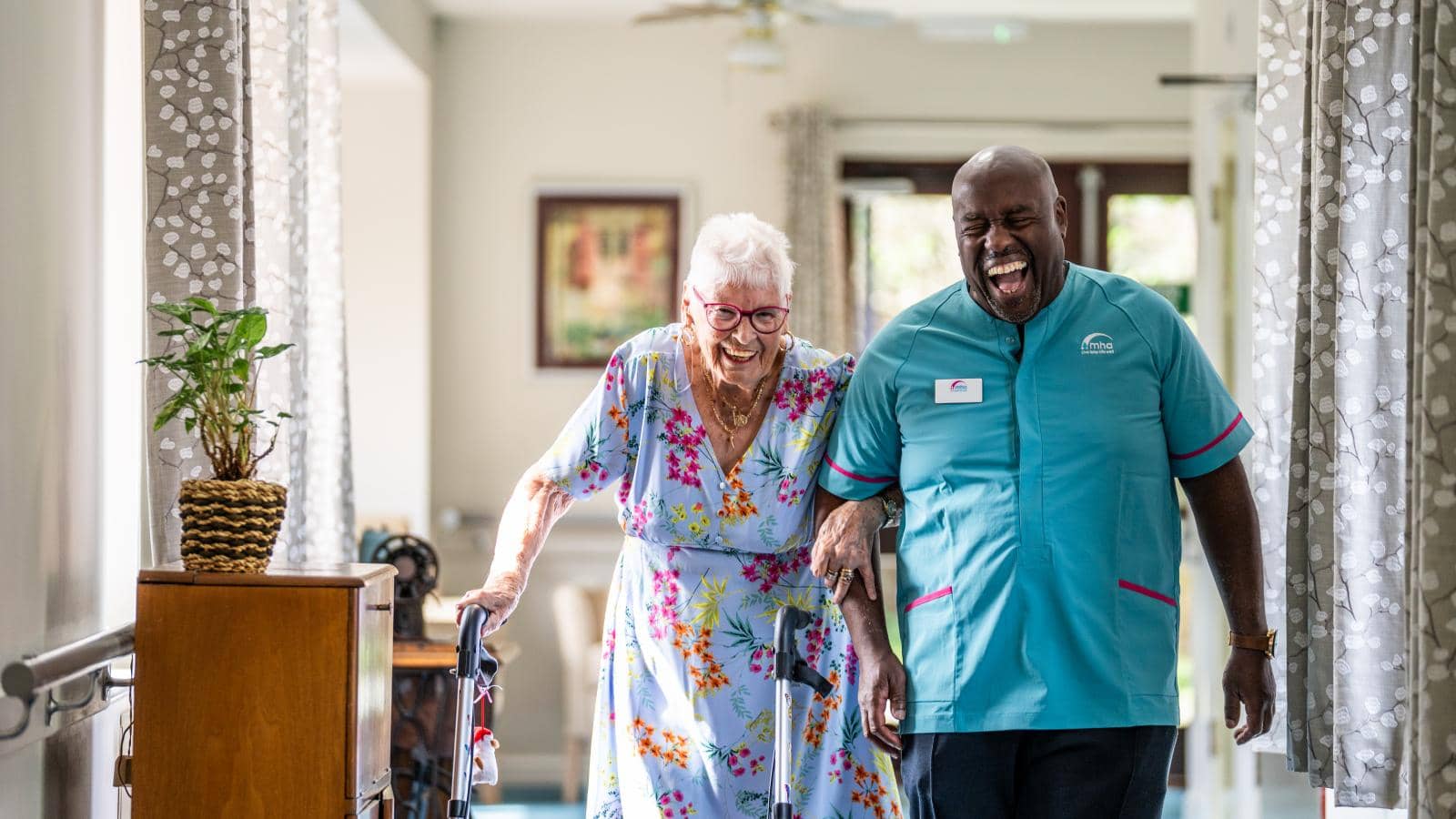 An elderly woman, wearing a colorful floral dress and glasses, is using a walker as she walks alongside a smiling caregiver. The caregiver, dressed in a light blue uniform, appears to be supporting her as they share a moment of laughter.