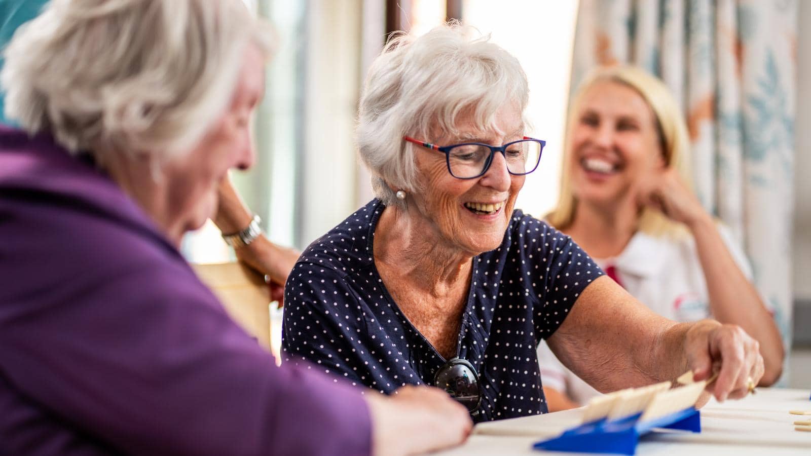 The image depicts a cheerful scene in a communal setting where a group of older women is engaged in a game or activity