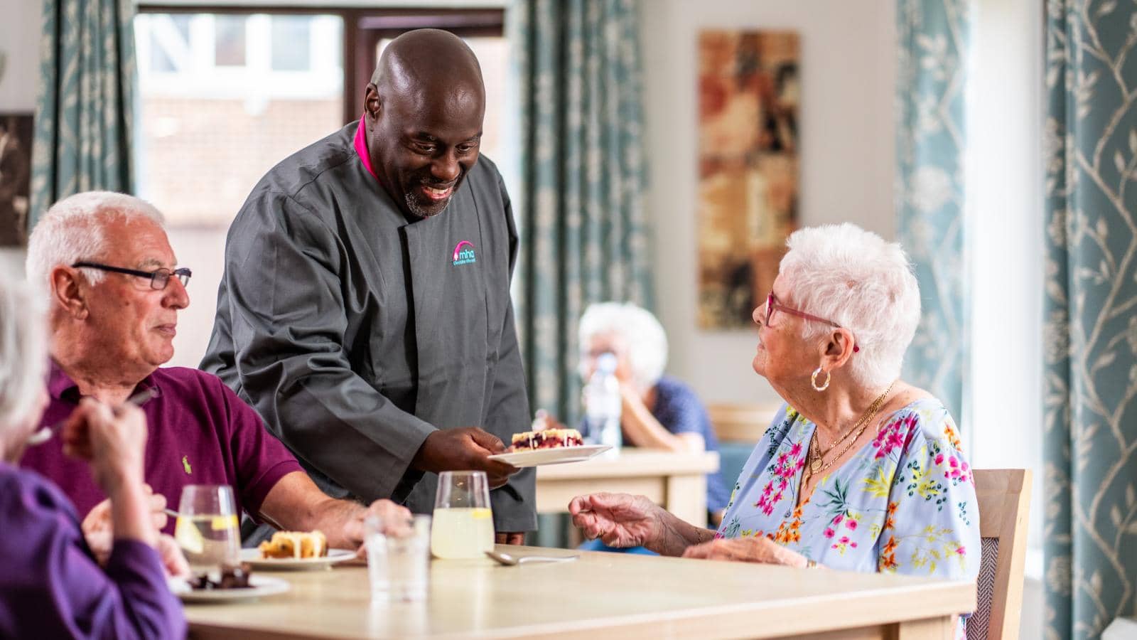 The image depicts a dining scene in a bright, inviting space. A cheerful male server, dressed in a dark chef's coat, is presenting a plate of food to an elderly woman with short, white hair, who is seated at a table.