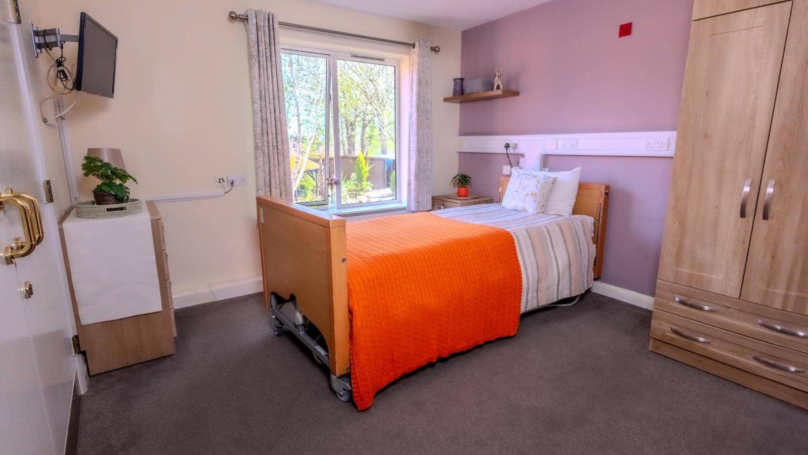 The image depicts a cozy bedroom designed for comfort and functionality. The room features a single bed with an orange blanket draped over it, complemented by light-colored bedding. A window allows natural light to fill the space