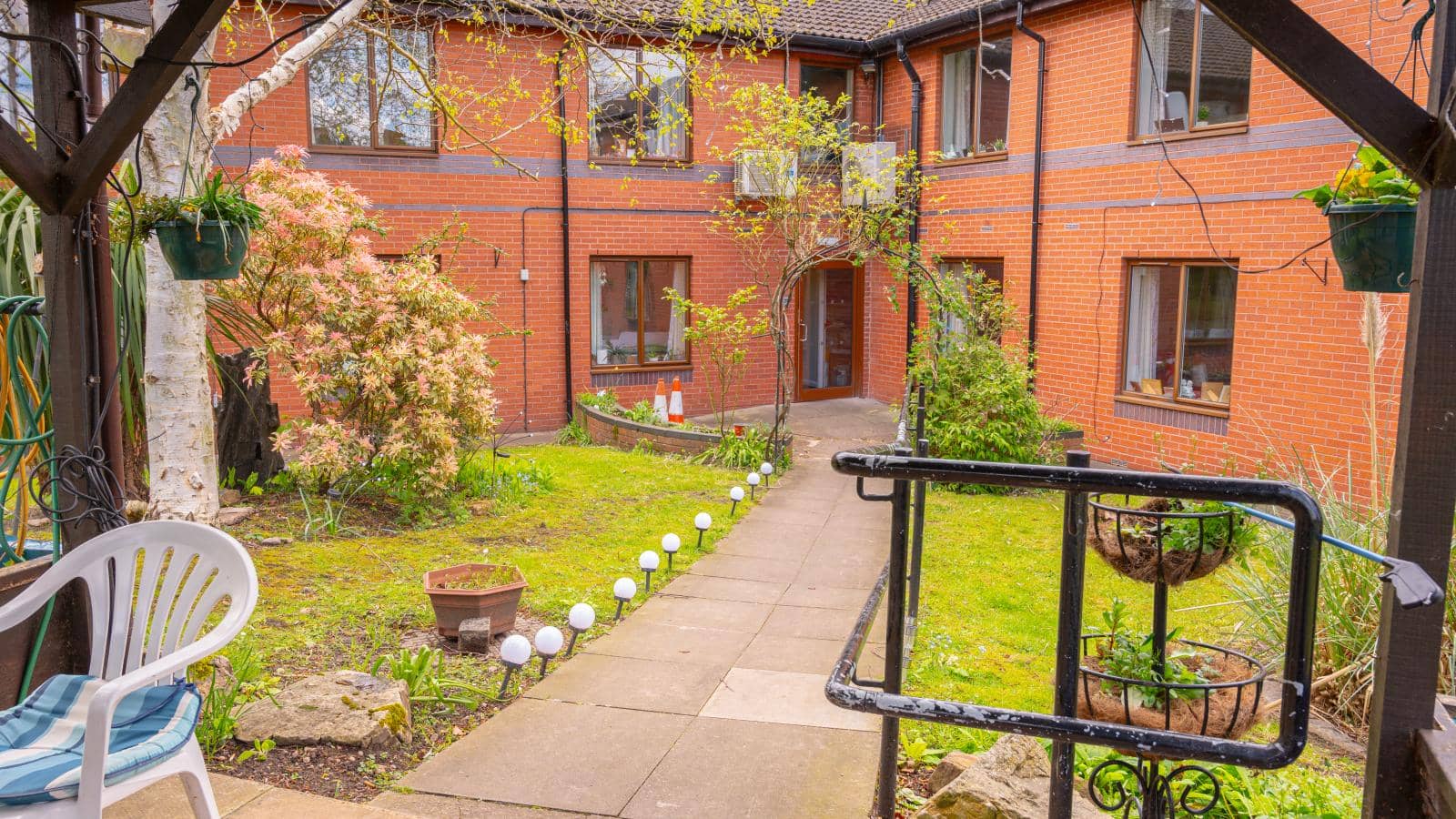 A charming courtyard surrounded by a brick building with several windows. The well-maintained garden has green grass, plants, and a flowering bush. 