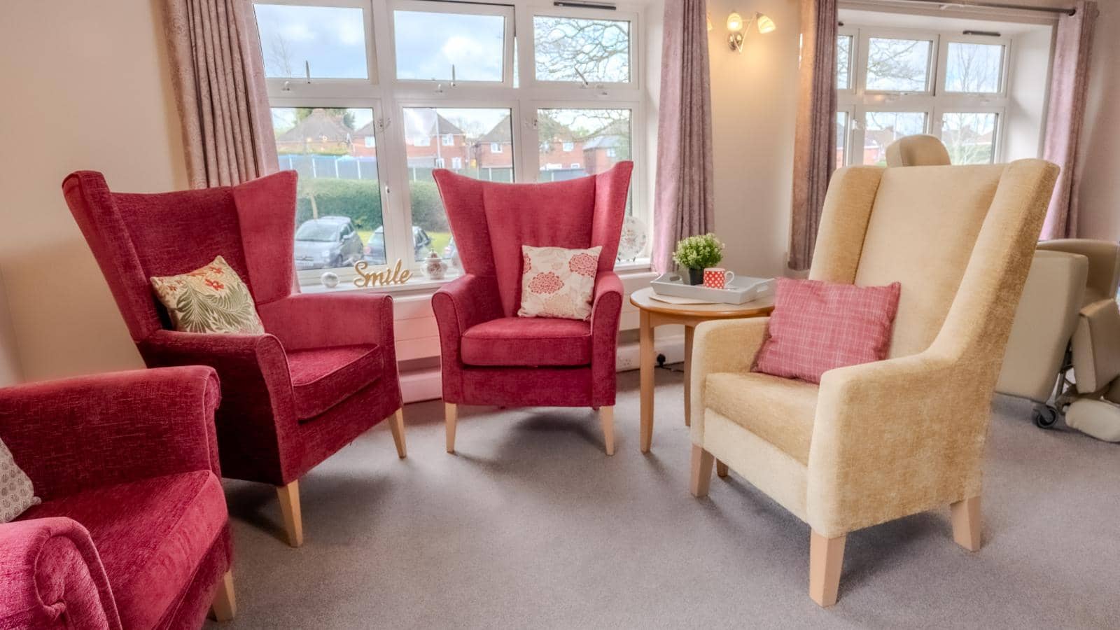 The image shows a cozy living space with two armchairs: one deep red and the other soft beige, both with high backs and wooden legs. Decorative cushions add warmth. Natural light streams through large windows with light curtains, showing greenery outside.