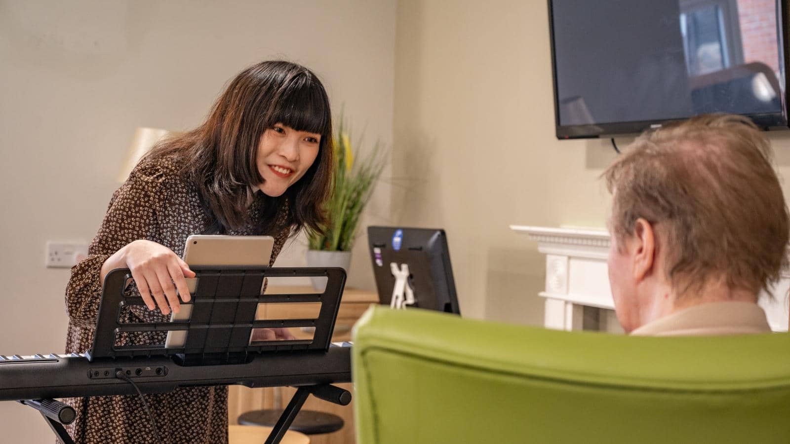 The image depicts a music therapist with long, dark hair, smiling as she interacts with an older man seated in a green chair. The setting is a cozy room with a light-colored wall.