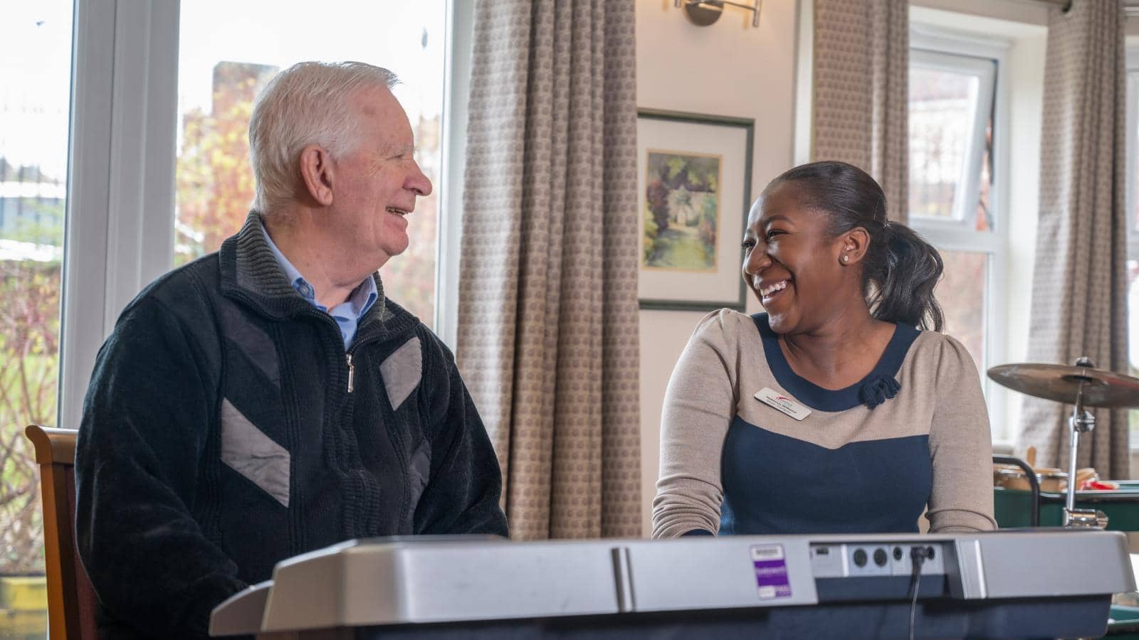 A resident and a music therapist sit side by side at an electronic keyboard, and the caregiver looks at him warmly, suggesting an engaging and positive interaction