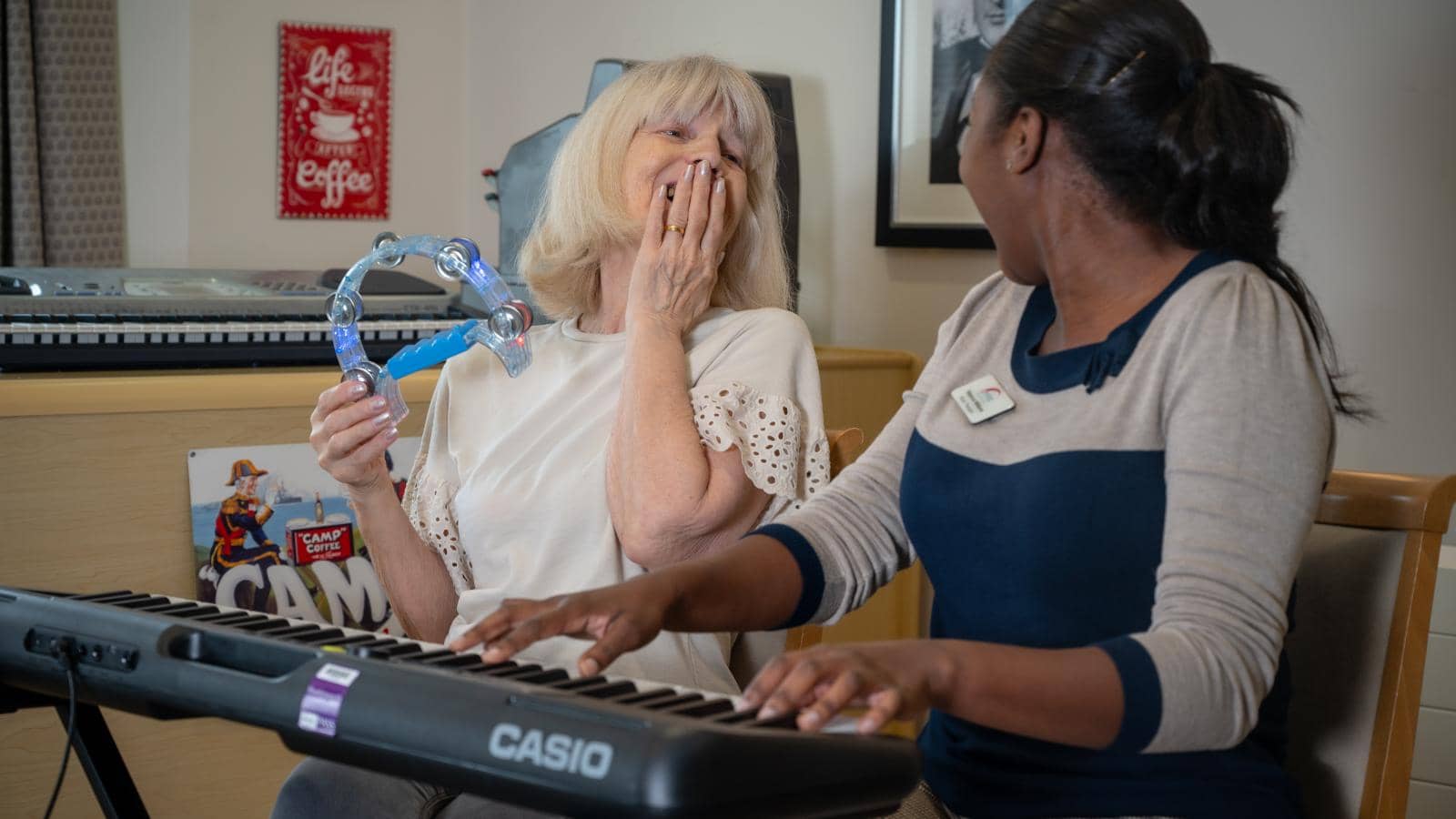 An elderly woman laughs while holding a tambourine during a music therapy session at Willowcroft. The music therapist plays the keyboard, sharing in the joyful moment. The background includes musical instruments and wall decorations