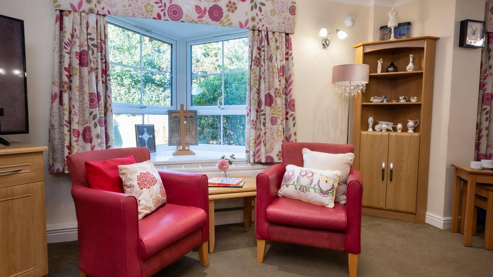 he image depicts a cozy living room with a warm and inviting atmosphere. The focal point is a pair of bright pink armchairs, each adorned with decorative cushions featuring floral and owl patterns