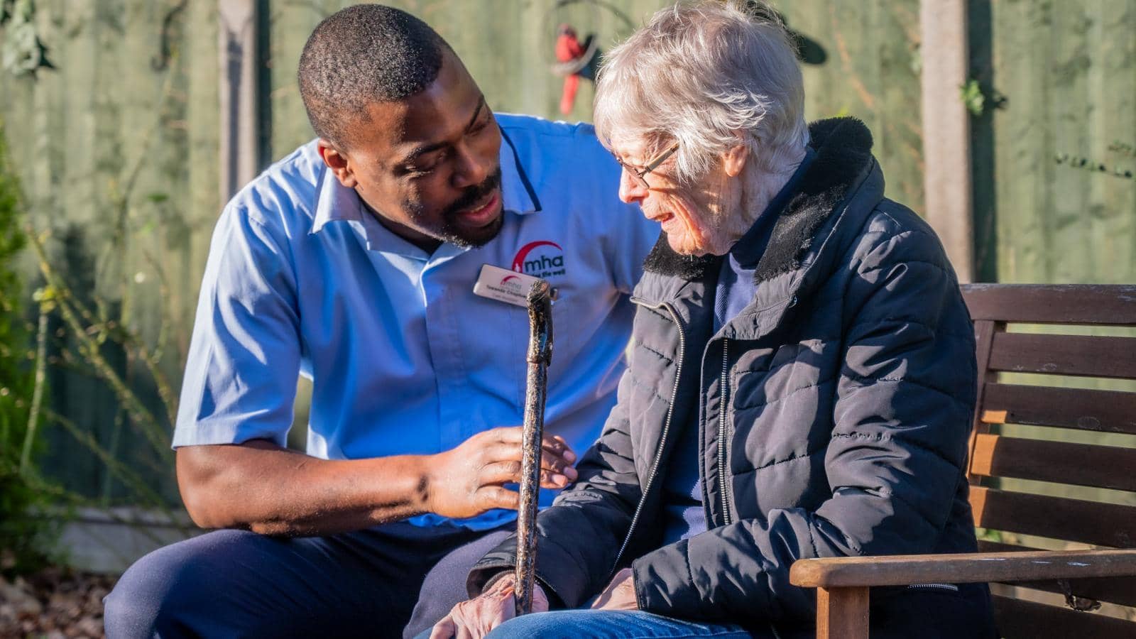 The image depicts a warm interaction between a caregiver and an elderly woman sitting on a bench in a garden setting. The caregiver, dressed in a light blue shirt, is attentively engaging with the woman, who appears to be in her 80s, wearing a dark jacket