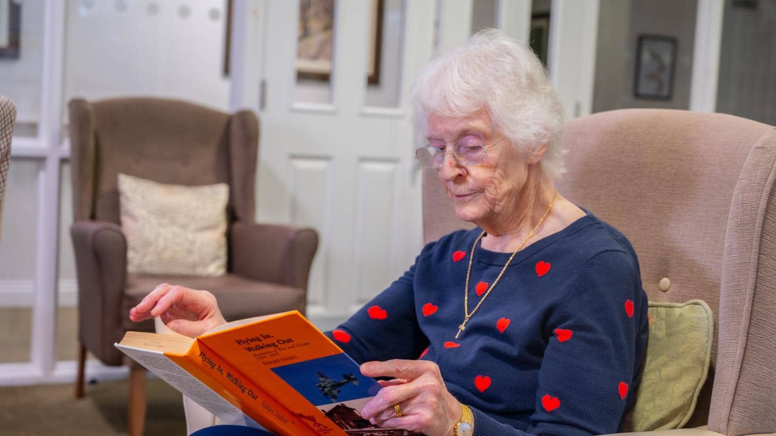 An elderly woman with white hair and glasses sits in a cozy chair, engrossed in reading a book with a bright orange cover. She wears a navy blue sweater with red heart patterns