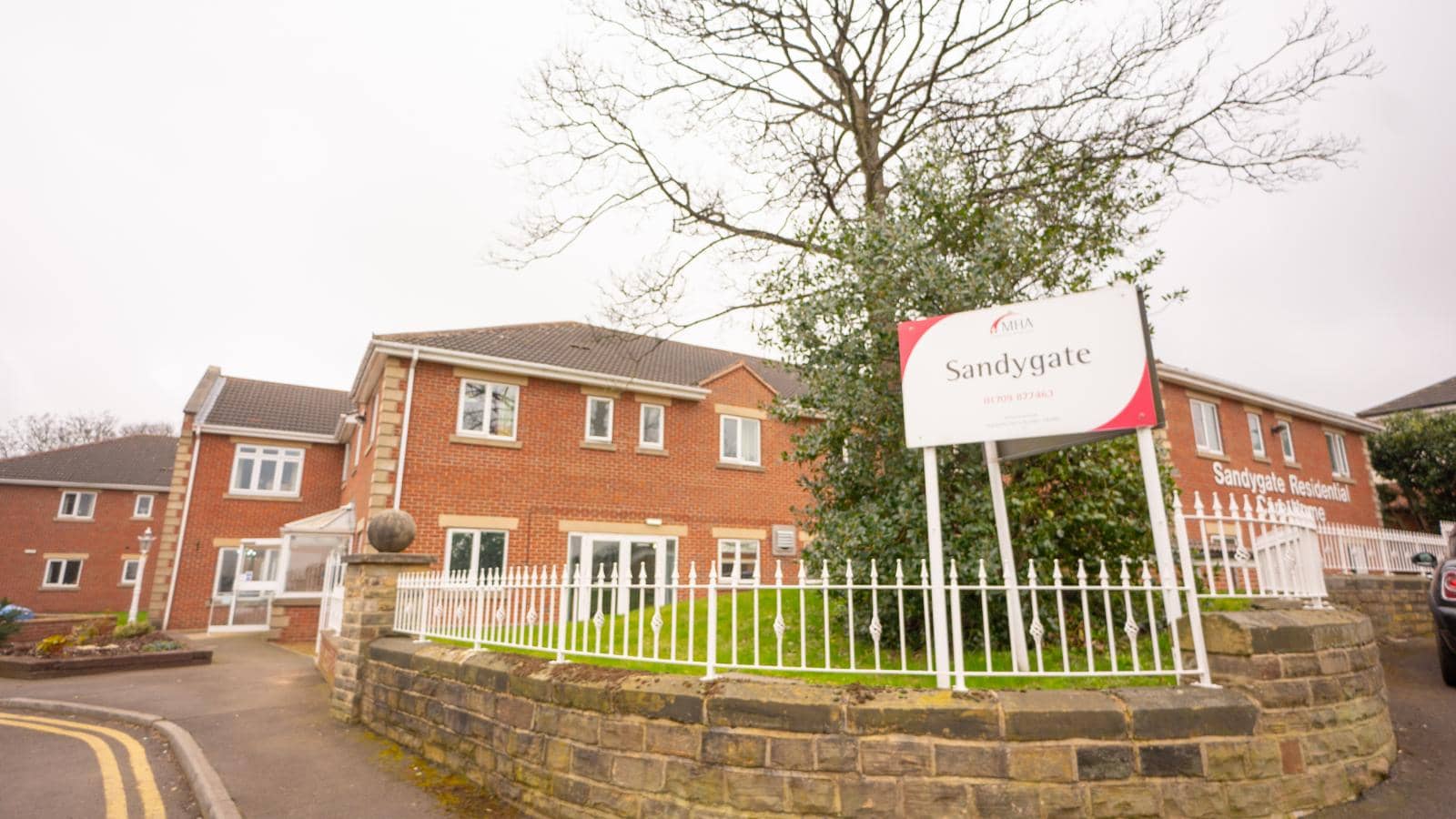 The image shows the exterior of Sandygate Residential, a red-brick care facility with white-framed windows. A sign near the entrance is surrounded by a white fence on a stone wall. The grounds have a lawn, tree, and shrubs.