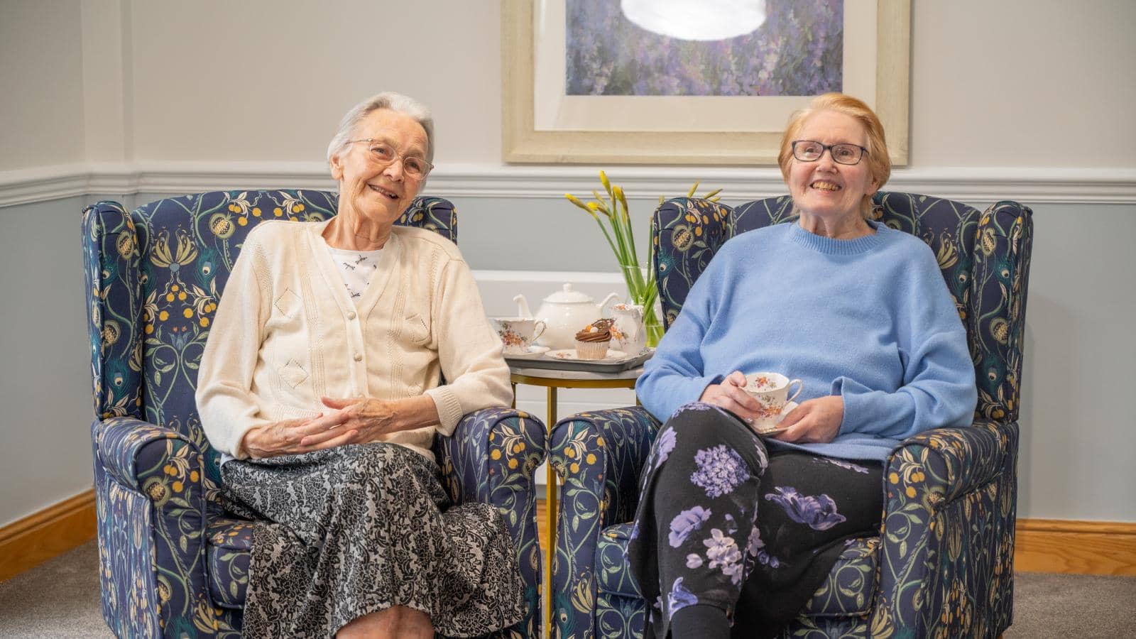 Two elderly women sit comfortably in patterned armchairs, smiling and enjoying each other's company. One wears a light cardigan over a white blouse, the other a light blue sweater and dark pan