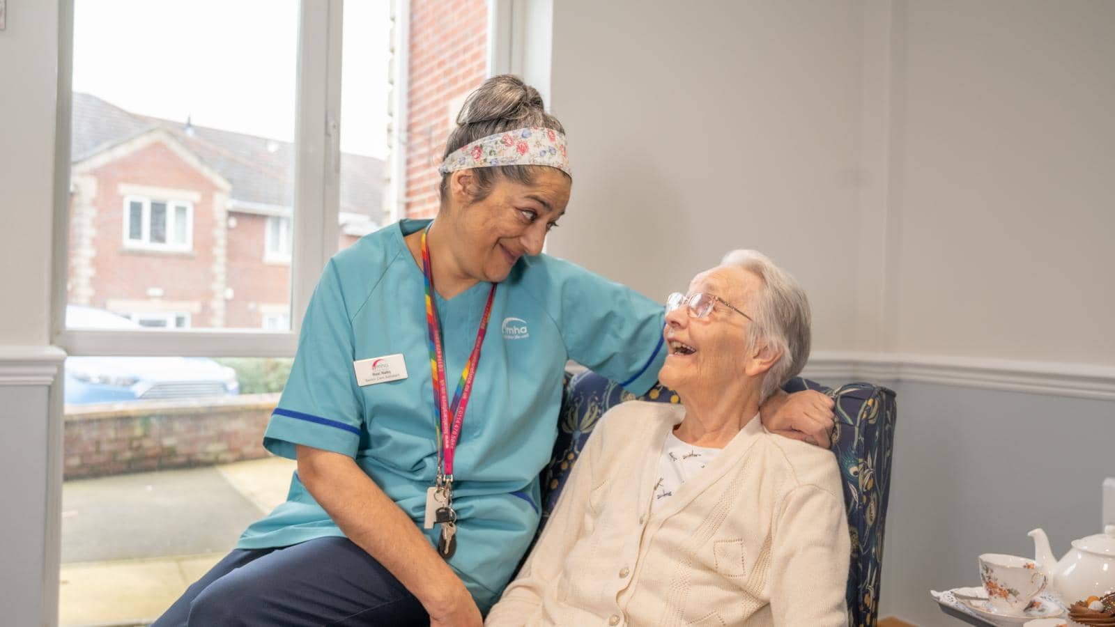  A healthcare worker in a blue uniform with a name badge and lanyard stands beside an elderly woman seated in a chair. The worker smiles warmly with her arm around the woman, who wears glasses and a light cardigan, looking up joyfully