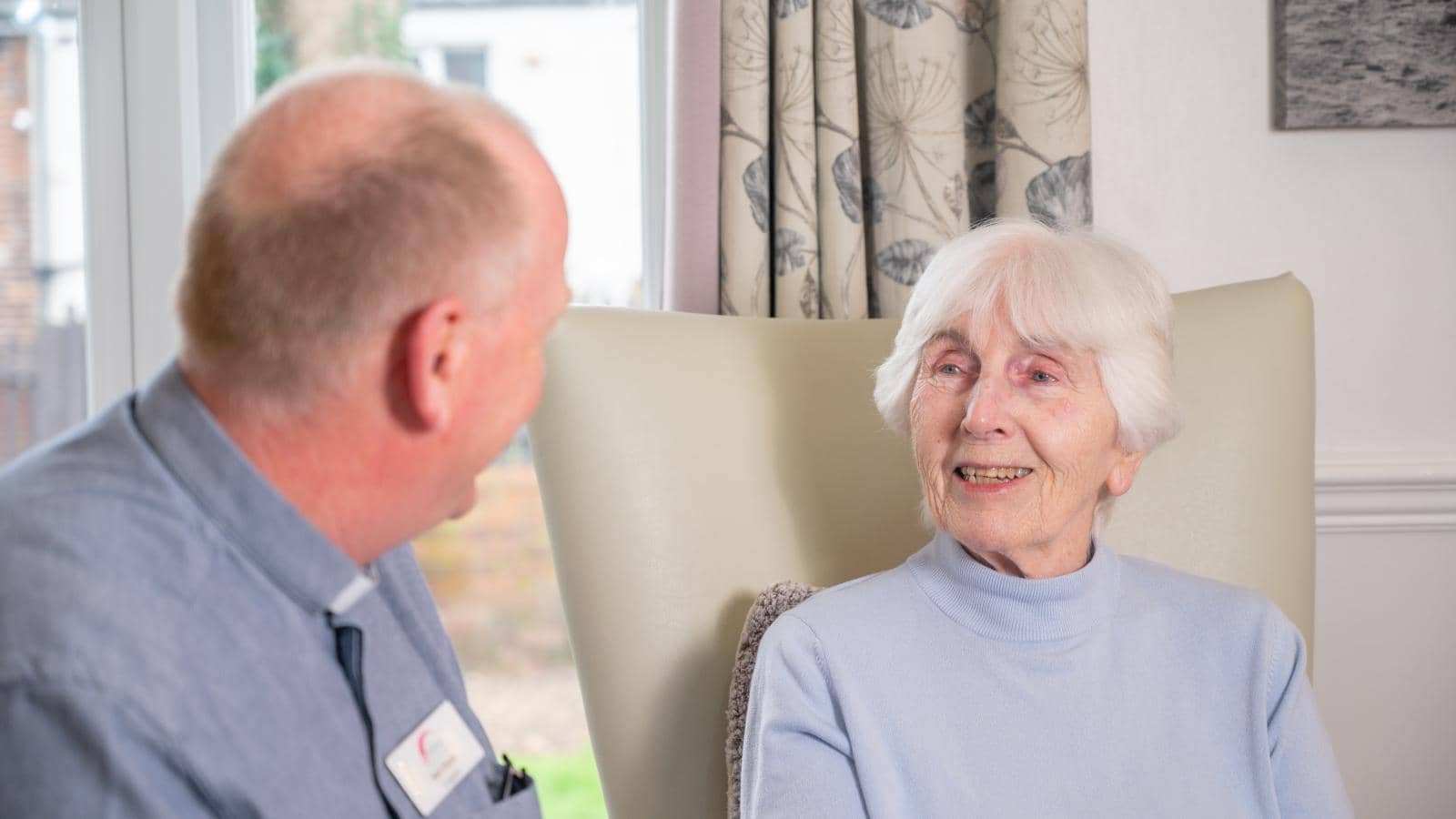 An elderly woman with short white hair sits in a light-colored chair, wearing a light blue sweater and smiling warmly. A male caregiver, dressed in a light blue shirt with a name badge, sits beside her, engaged in conversation