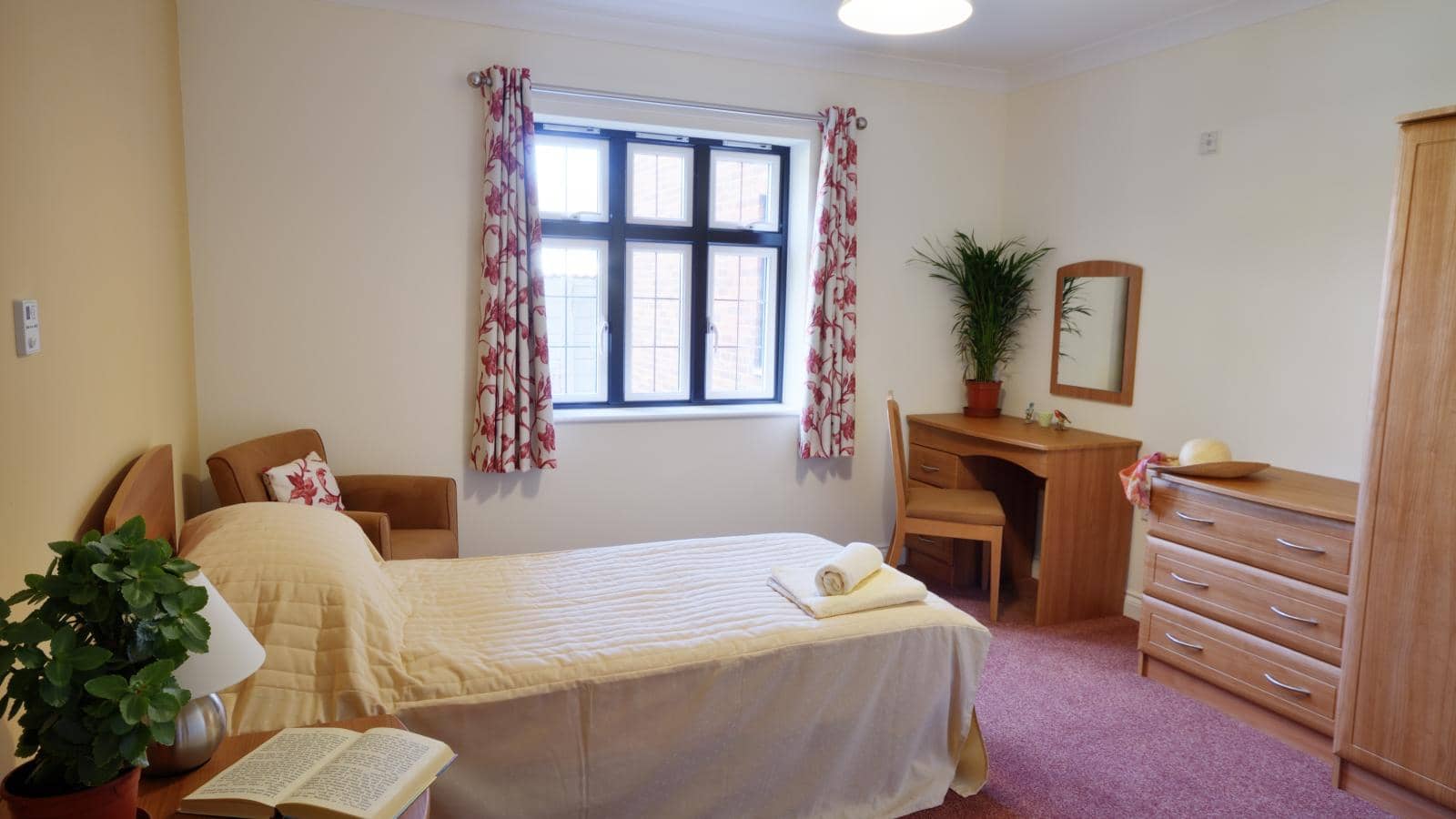 Two women interact in a cozy room. The caregiver, smiling, is seated on the left with long hair. The elderly woman on the right has short hair, glasses, and holds a book. The room features large windows, curtains, and comfortable furniture.