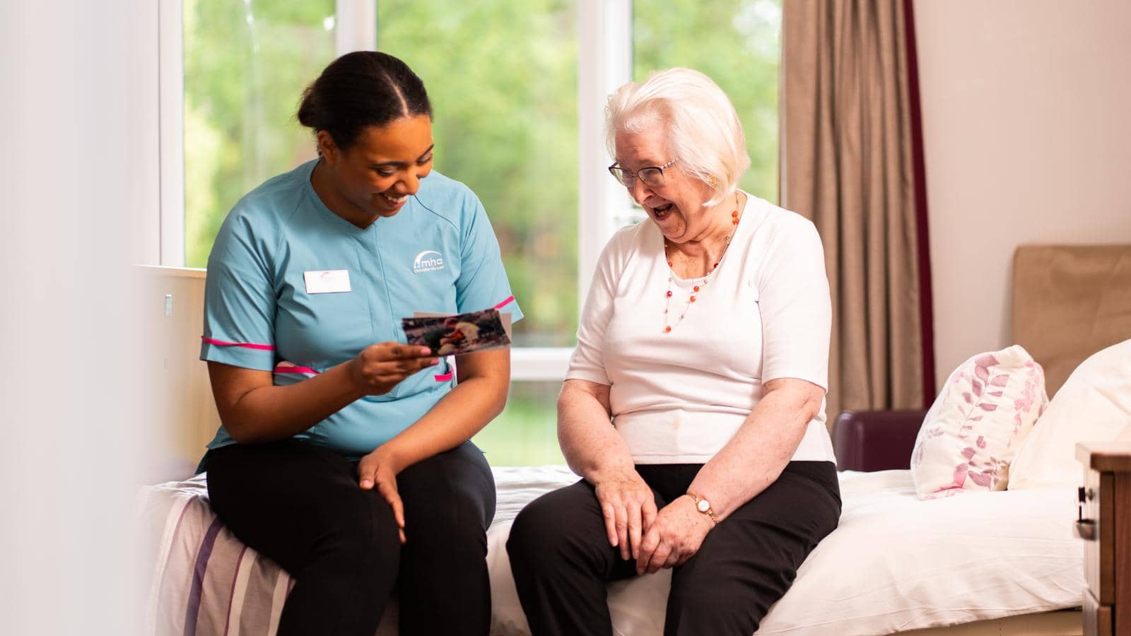 Two women share a warm interaction in a well-lit room. A caregiver in a light blue uniform with a name tag sits on the edge of a bed, holding a photograph for the elderly woman with white hair, glasses, and a white top. 