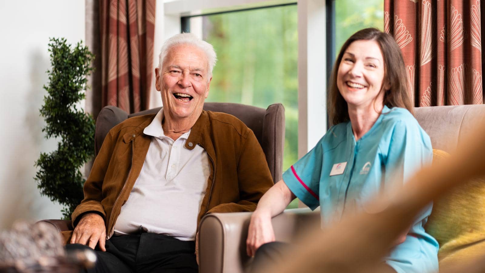 An elderly man and a woman sit in a comfortable, well-lit room. The man, in a brown jacket over a white shirt, smiles in an armchair. The woman, a caregiver smiles warmly. A window with curtains and a potted plant are visible.