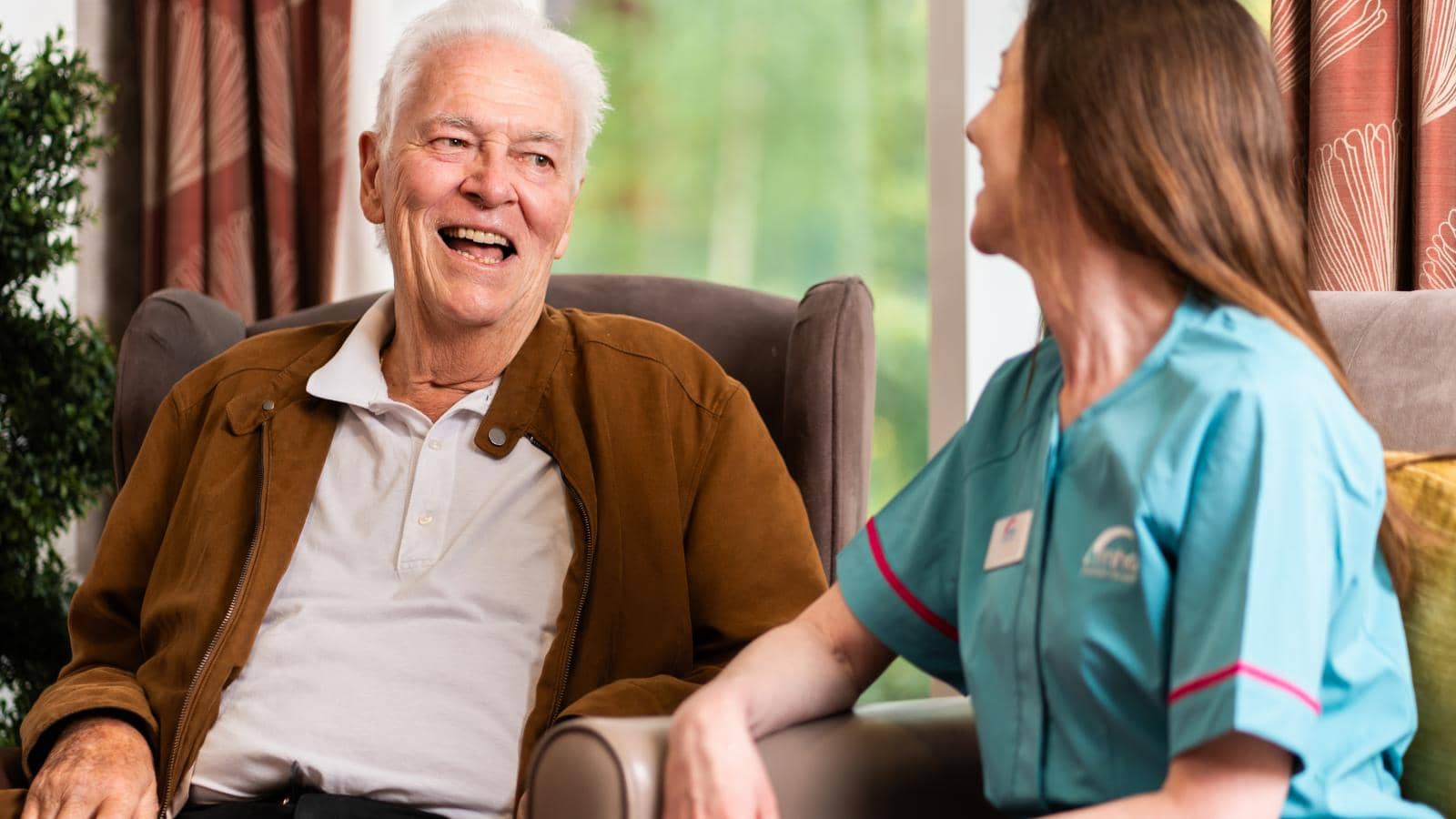 An elderly man with white hair, wearing a brown jacket over a white shirt, sits in a well-lit room, smiling warmly in conversation with a caregiver. The caregiver, in a light blue uniform with pink trim, smiles back.