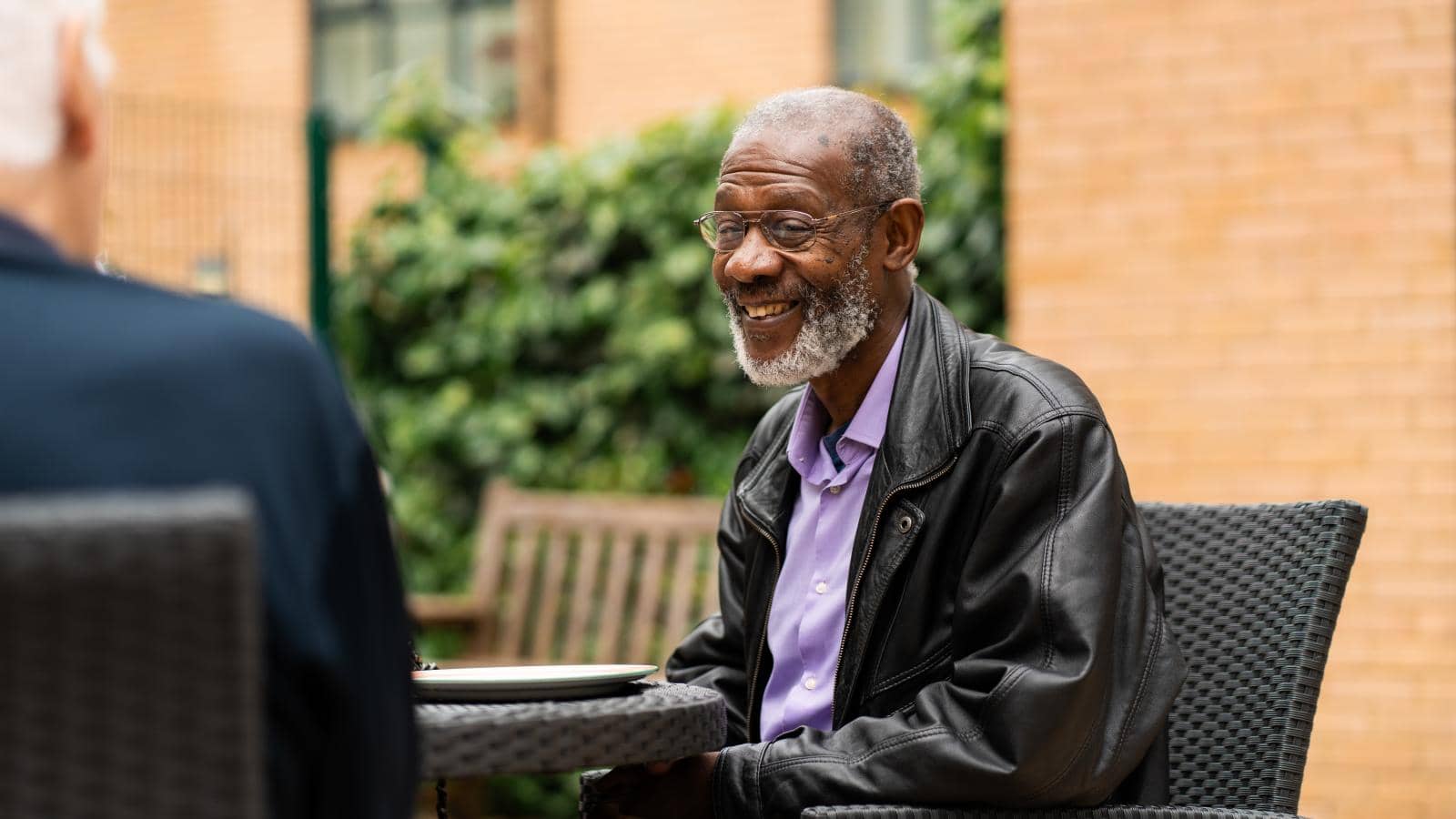  An elderly man sits outdoors at a table, smiling warmly during a conversation with someone whose back faces the camera. He wears a black leather jacket over a light purple shirt and glasses. 