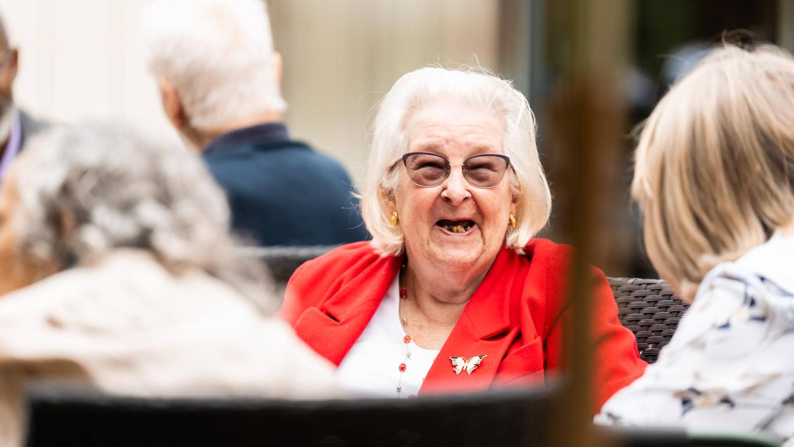 An elderly woman with white hair, glasses, and a bright red blazer with a butterfly brooch sits outdoors, laughing or smiling at a social gathering. Other people with gray and light-colored hair are visible in the foreground. 