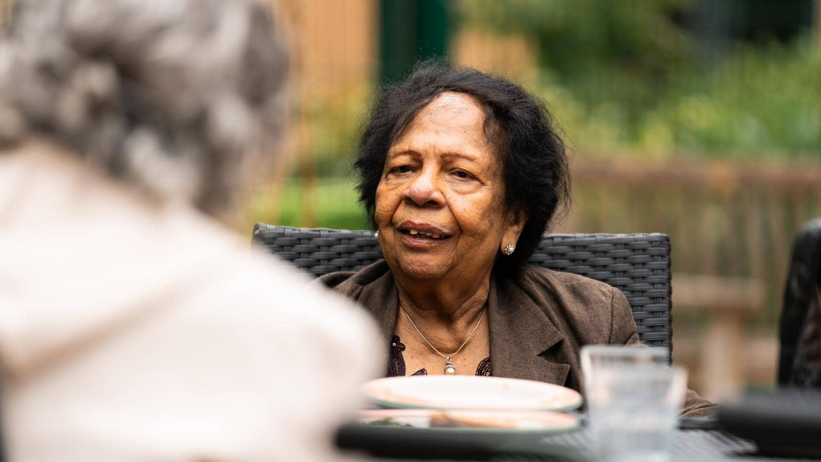  An elderly man sits outdoors at a table, smiling warmly during a conversation with someone whose back faces the camera. He wears a black leather jacket over a light purple shirt and glasses. 