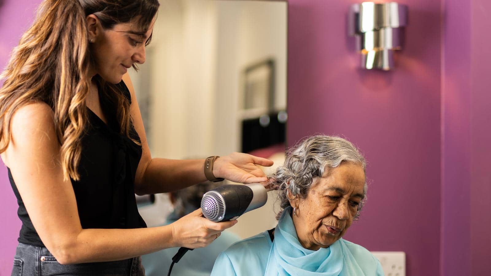 An elderly woman with gray hair sits in a salon, wearing a light blue outfit and matching scarf, eyes closed and relaxed. A hairstylist with long brown hair, in a black top and dark jeans, uses a hairdryer.