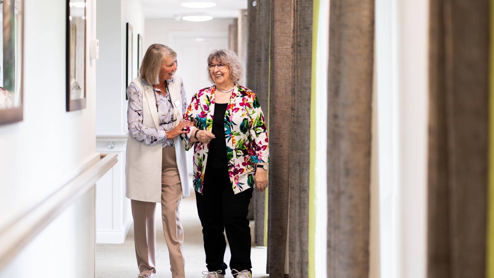 Two women walk down a well-lit hallway, smiling and conversing. The woman on the left, with shoulder-length gray hair, holds the arm of the other woman. The woman on the right, with short gray hair and glasses, smiles as they talk.