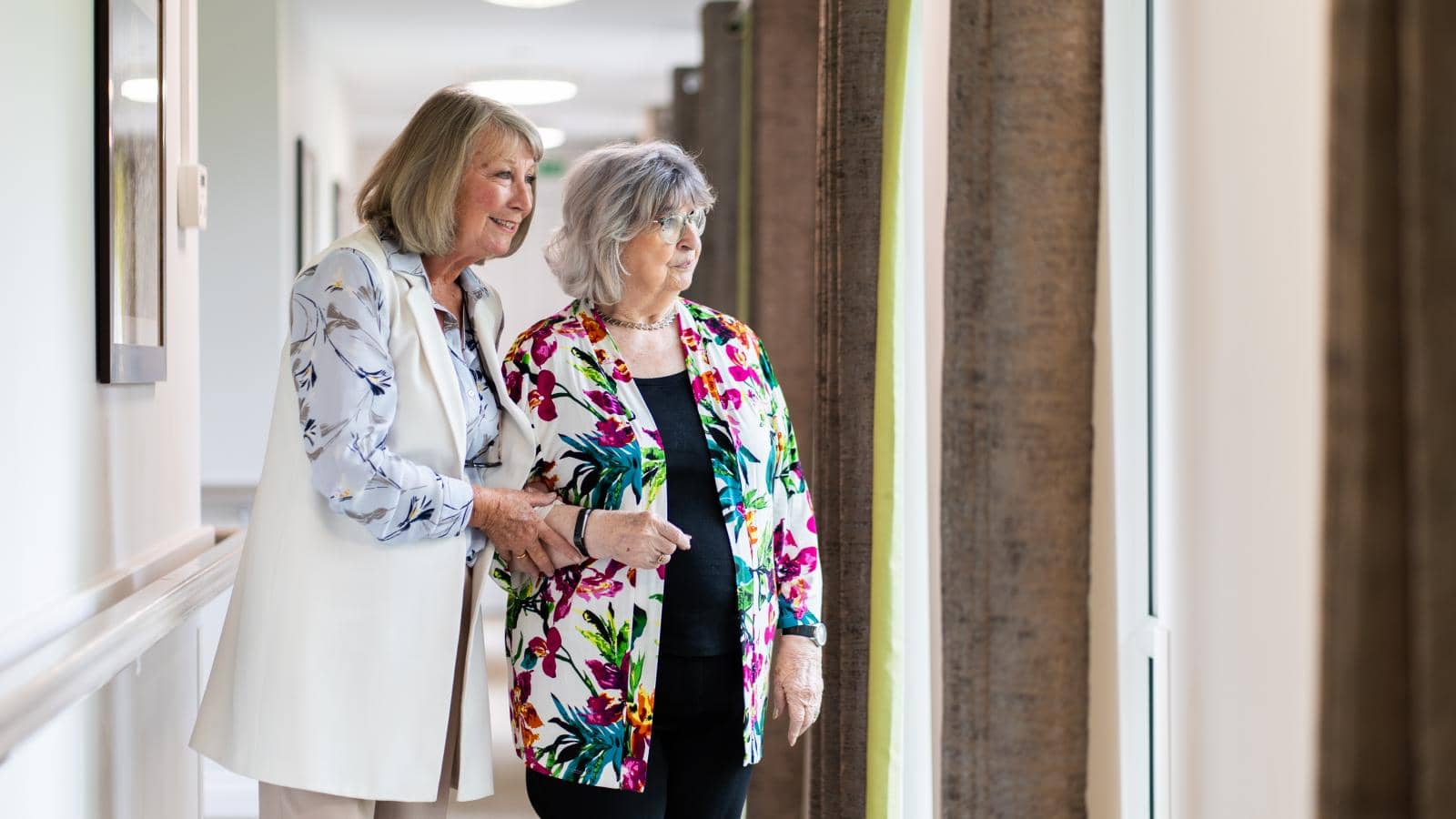 Two elderly women stand side by side, looking out of a window. The woman on the left, with shoulder-length blonde hair, gently holds the arm of the woman on the right, who has short grey hair and glasses.