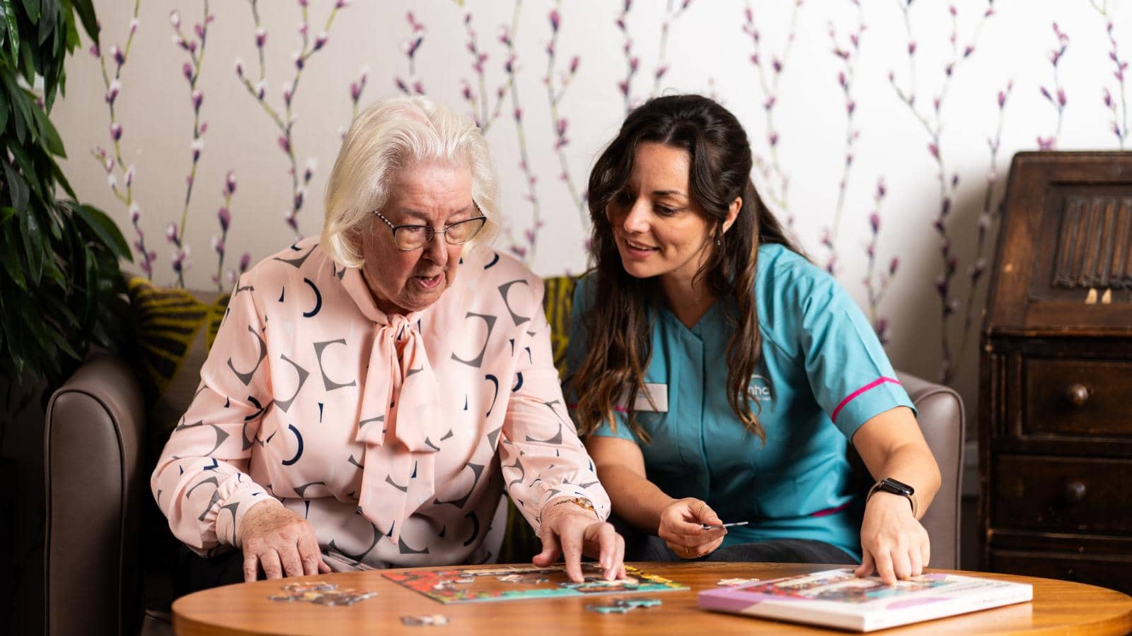 Two women sit at a round wooden table, assembling a jigsaw puzzle. The elderly woman, with white hair and glasses, wears a light pink blouse with a bow and appears focused. The younger woman, in a teal uniform, smiles while assisting.