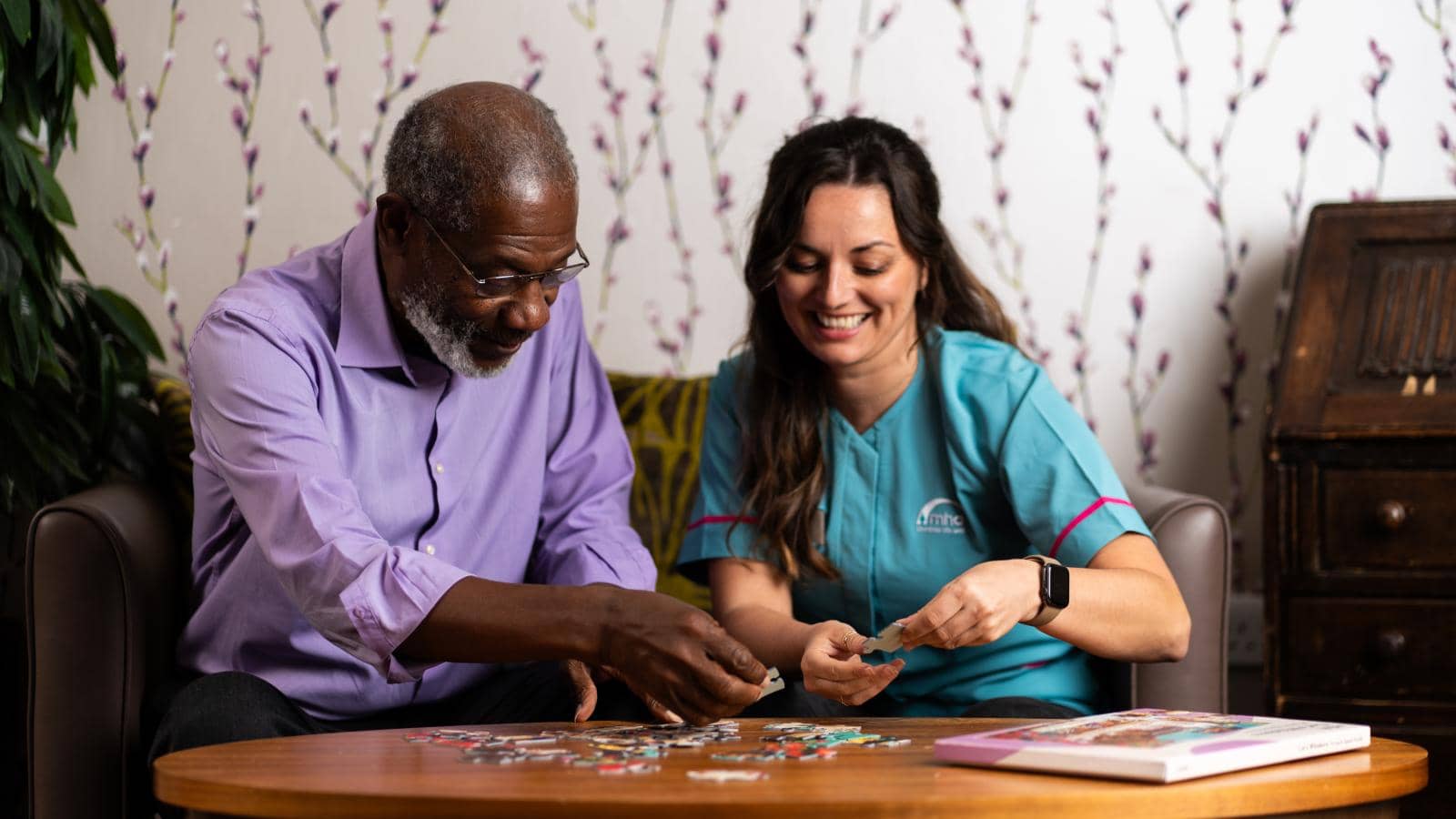  An elderly man in a light purple shirt and a younger woman in a teal uniform sit at a wooden table, working on a jigsaw puzzle. He reaches for a piece while she smiles, engaged in the activity.