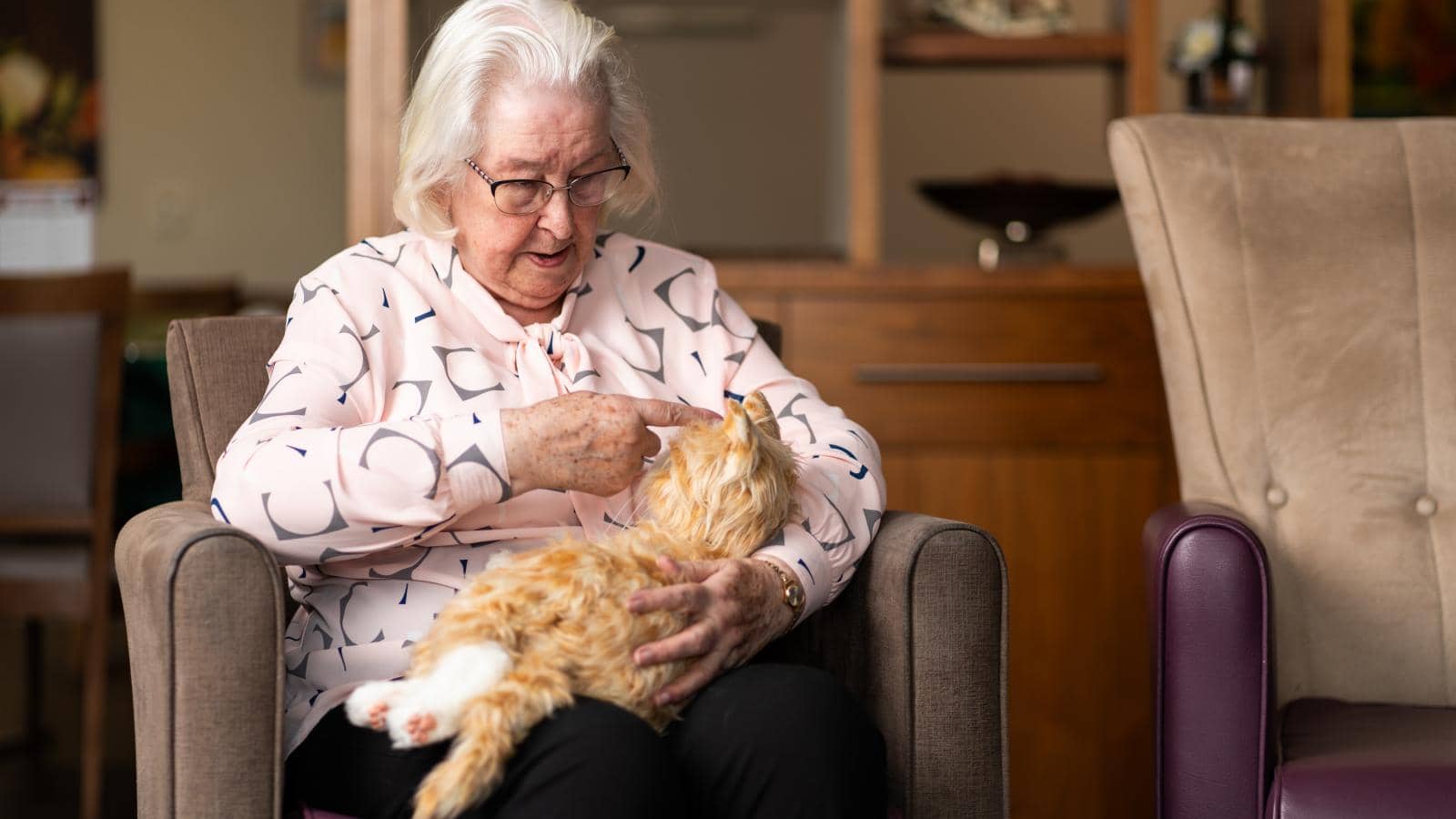 An content, elderly woman sitting in a comfortable chair, gently petting a small, ginger robotic cat that is resting on her lap, in a cozy, well-lit room with wooden furniture and soft, neutral-colored decor.