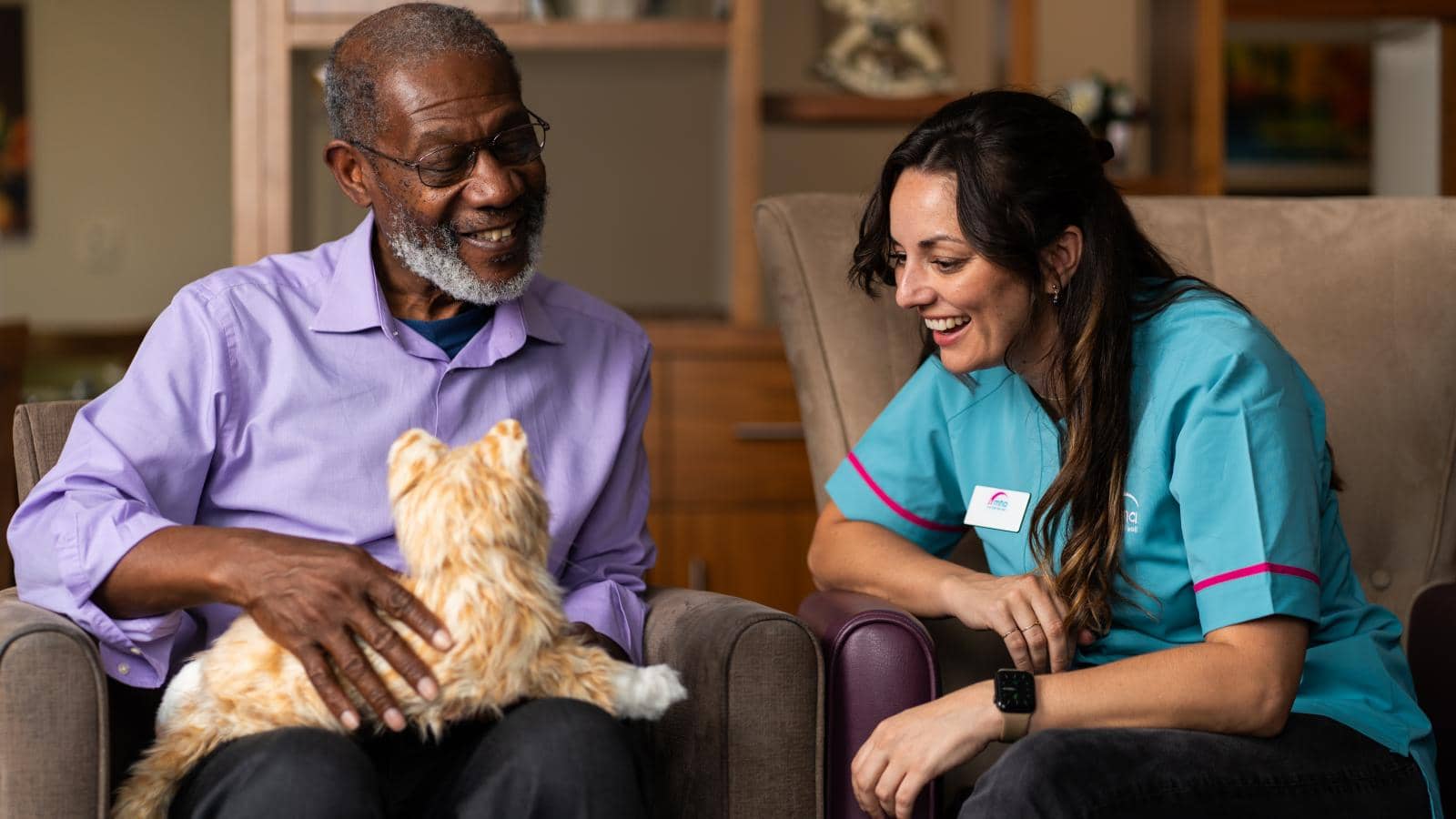 An elderly man in a light purple shirt sits in a chair, holding a lifelike robotic cat, a comforting aid for dementia, and smiling. A younger woman in a teal uniform with pink trim sits beside him, also smiling.