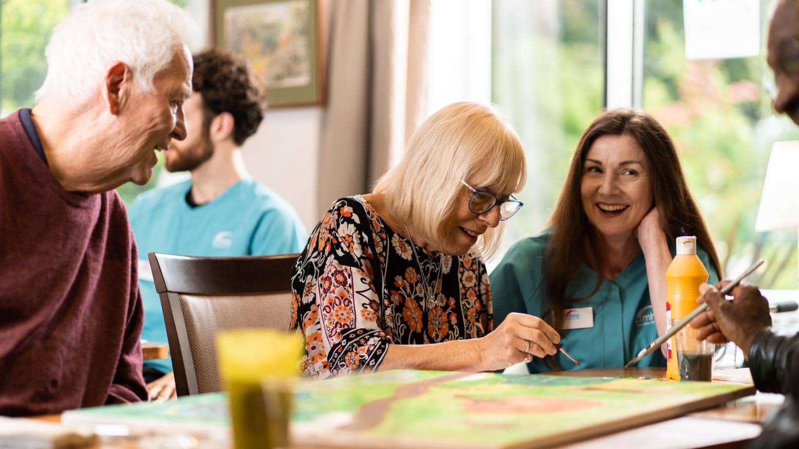  The image shows a lively scene in a community or care center, where several people are seated around a table, participating in a painting activity. In the foreground, an elderly woman with blonde hair and glasses focuses on her colorful painting