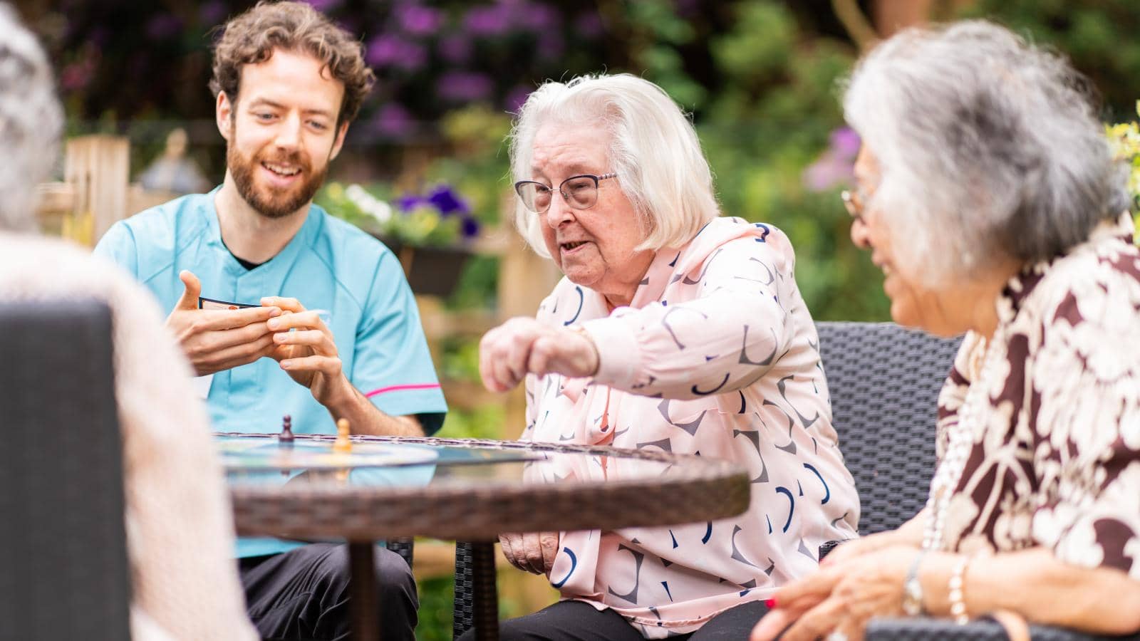 A group of elderly individuals and a caregiver sit around a table outdoors in a garden or patio area with greenery and flowers in the background. The caregiver, in a light blue uniform, smiles and interacts with the group as they engage in conversation 