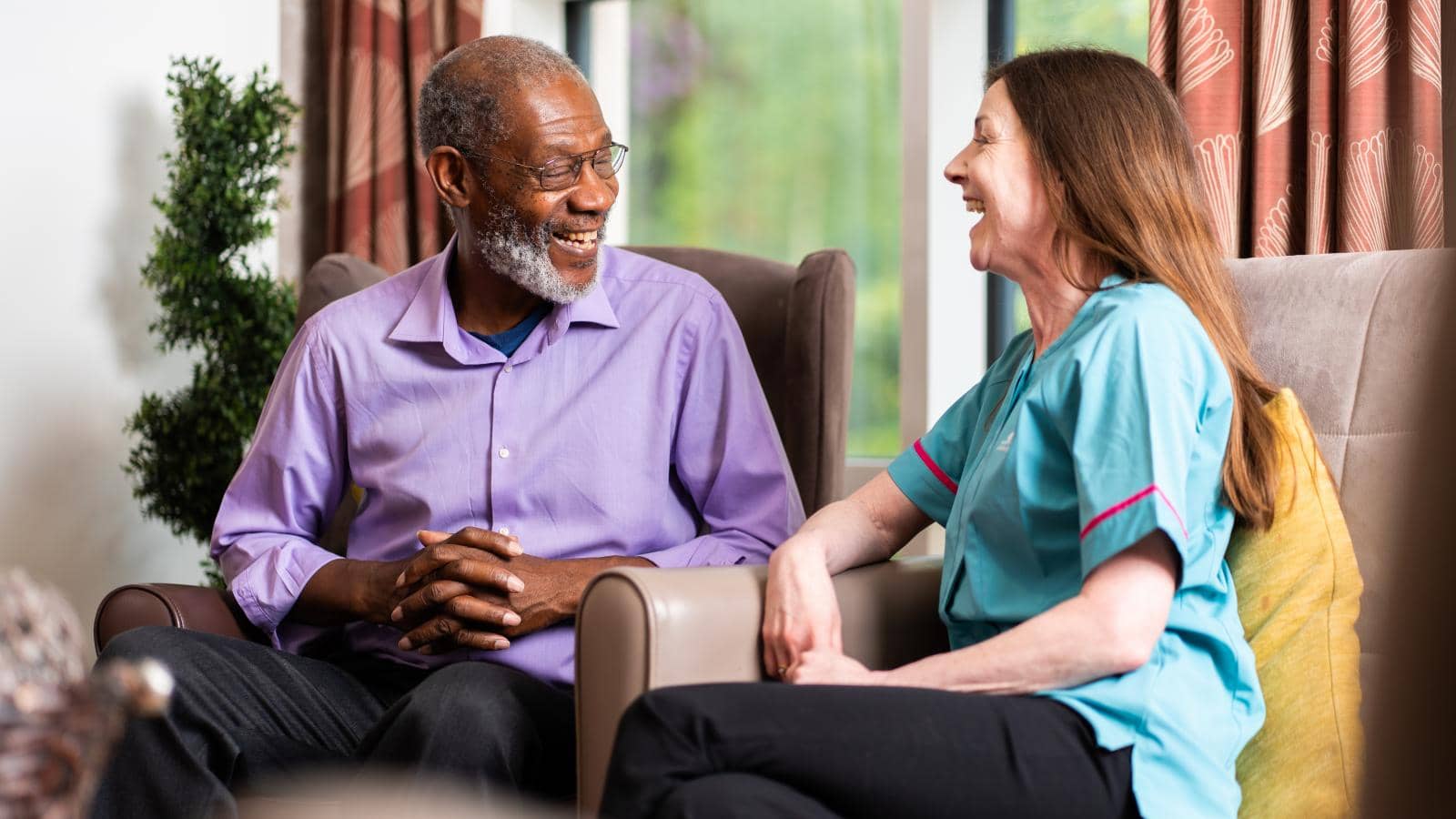 Two people sit in a comfortable, well-lit room. The older man on the left, with gray hair, a beard, and glasses, smiles while engaged in conversation. The woman on the right, with long brown hair in a light blue scrub top, listens attentively