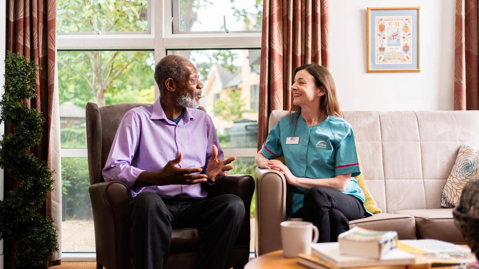 An elderly man in a light purple shirt sits in an armchair, gesturing during a lively conversation with a caregiver in a teal uniform with a name tag. She smiles warmly from a nearby sofa. 