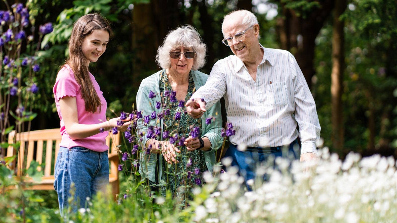 Three people stand together in a garden among blooming flowers. An elderly man points towards purple flowers, while an elderly woman in sunglasses and a younger woman with long brown hair observe the scene. 