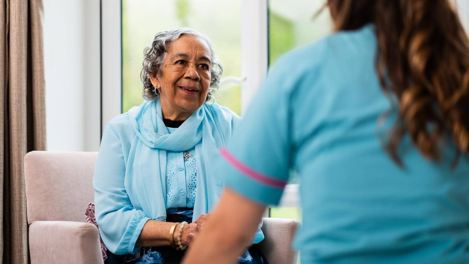 An elderly woman with curly gray hair sits in a chair, wearing a light blue scarf over a matching blouse and floral skirt, smiling as she talks with a caregiver in a light blue uniform.