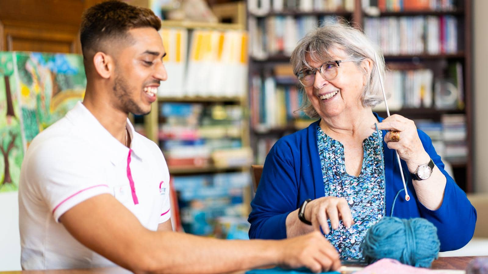 A young man and older woman sit at a table. The man in a white polo smiles at her. She, with gray hair, glasses, and a blue cardigan, holds knitting needles. The table has yarns. The cozy room with bookshelves adds a warm atmosphere.