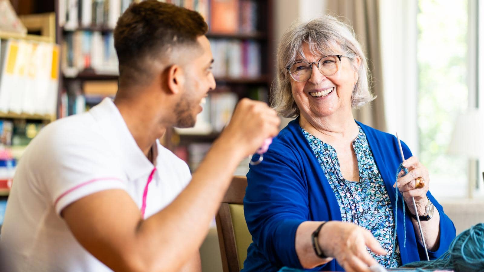 An elderly woman with gray hair and glasses smiles warmly, holding knitting needles, suggesting she's engaged in knitting. A younger man with short dark hair sits beside her, also smiling as they converse