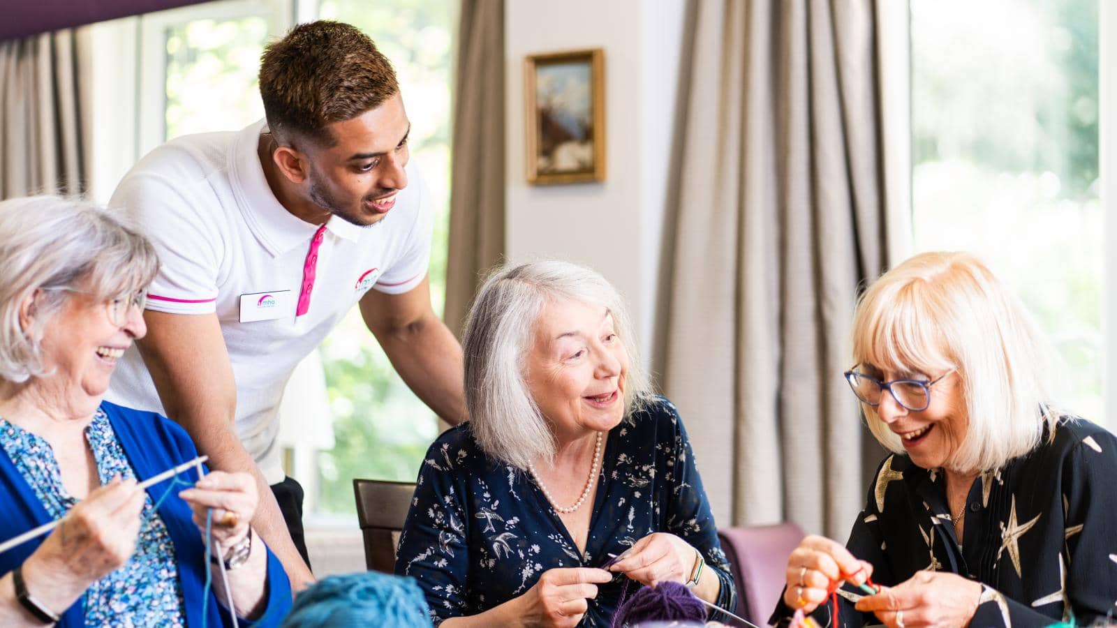 A cheerful group of elderly women sits around a table, engaged in knitting with colorful yarn, smiling and chatting together. A young man stands nearby, leaning in to engage with them