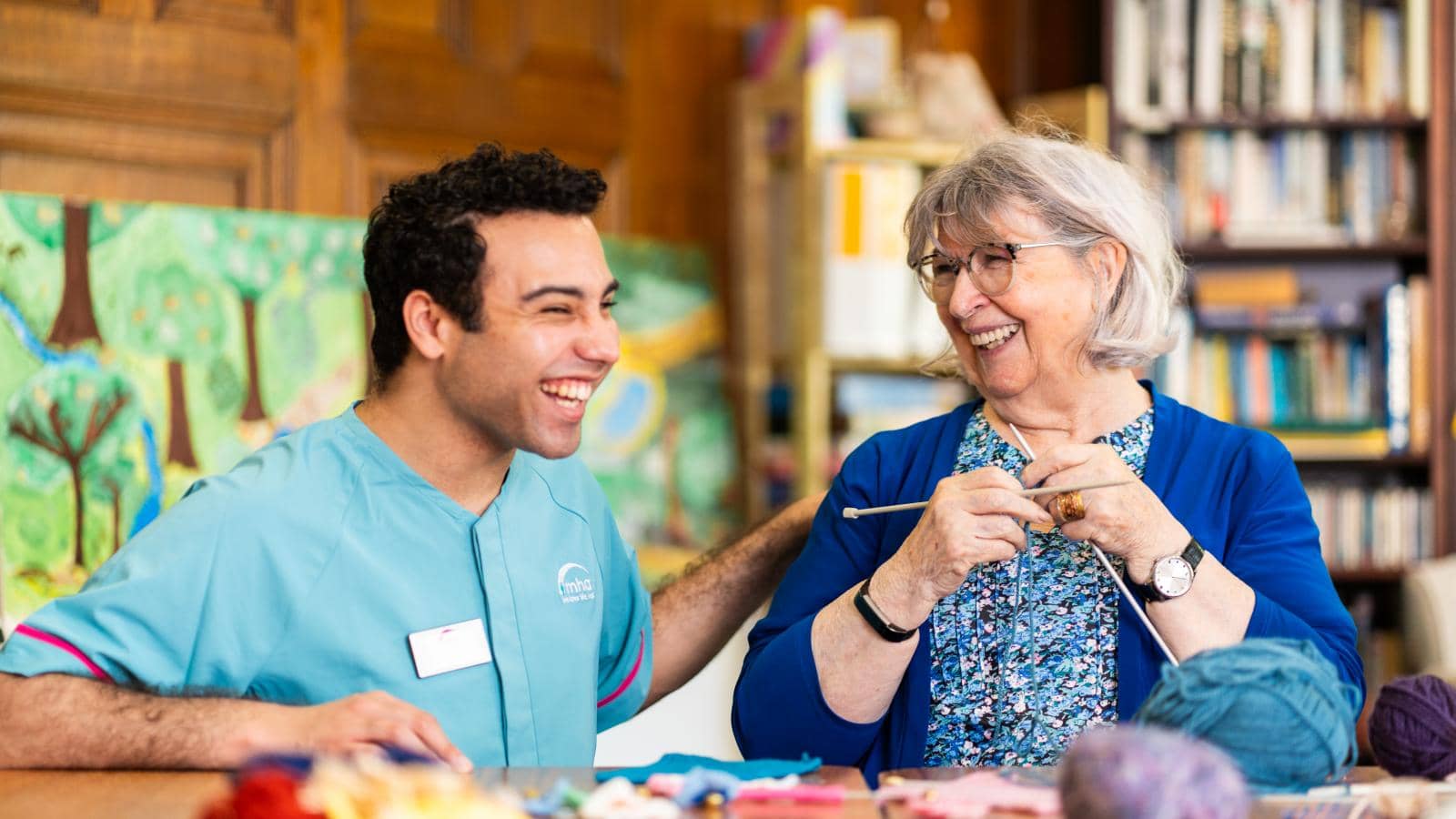 A man in a light blue uniform with pink accents, smiling broadly, sits on the left, engaged in conversation with an older woman with gray hair and glasses. She wears a blue cardigan over a patterned blouse and holds knitting needles.