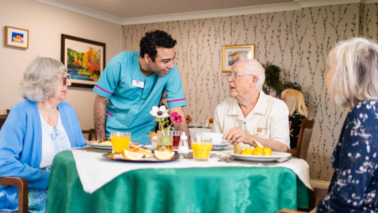 Four elderly individuals sit around a table with a green tablecloth, enjoying a meal together in a dining area. A caregiver stands nearby, engaging attentively with them. The table is set with food, juice, and a vase of flowers